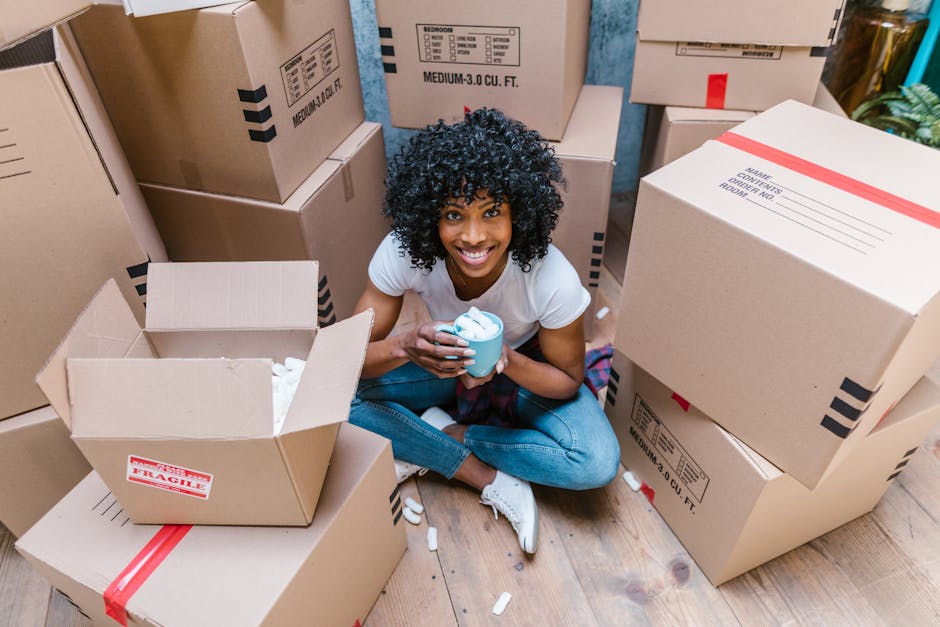 A young woman with curly black hair and a bright smile is sitting on a wooden floor surrounded by stacked cardboard boxes, some labeled as fragile, in a room. She is holding a blue mug filled with a b