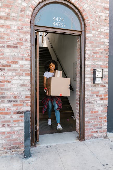 A young woman with curly black hair and a bright smile is sitting on a wooden floor surrounded by stacked cardboard boxes, some labeled as fragile, in a room. She is holding a blue mug filled with a b
