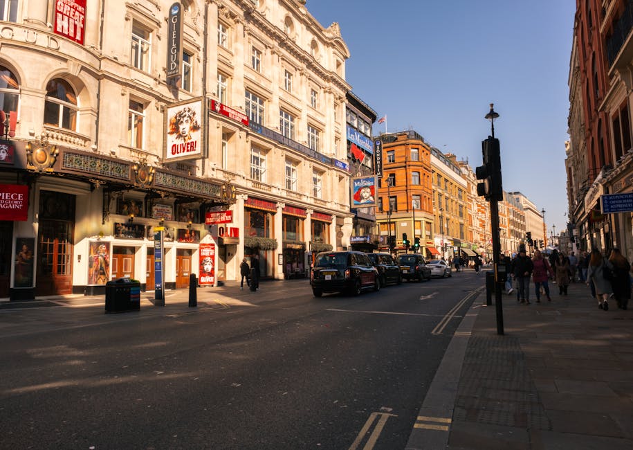 A busy street scene in an urban area with historic multi-storey buildings featuring detailed facades and large windows, located along Fulham Broadway. The street is lined with shops and commercial establishments, including a theatre with posters and signage visible on the building exteriors. Several pedestrians are walking on the sidewalk, some carrying shopping bags or engaged in conversation, while vehicles including cars and a van are driving along the road. A traffic light pole with illuminated signals is situated near the edge of the pavement, which has tactile paving for accessibility. The scene is captured during daylight with clear blue skies, and the lighting casts shadows on the street and building facades, reflecting a typical environment for home relocation and furniture transport logistics involving [COMPANY_NAME] and its removals services near Fulham station.