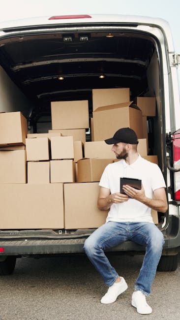 A man wearing a white t-shirt, blue jeans, and a black cap sits on the ground beside the open rear doors of a large delivery van, which is filled with numerous cardboard boxes of varying sizes stacked neatly inside. The boxes are sealed with packing tape and are arranged to maximize space within the van. Some boxes are positioned on top of others, and a few are placed near the doorway to facilitate loading or unloading. The man is holding and looking at a tablet device, possibly reviewing inventory or scheduling. The scene appears to be outdoors, with the van parked on a paved surface, capturing a moment during home relocation or furniture transport as part of a prompt for house removals. This image exemplifies packing and loading processes typical for professional removals services like those provided by Fulham Man and Van, emphasizing careful handling and organization during moving and packing tasks.