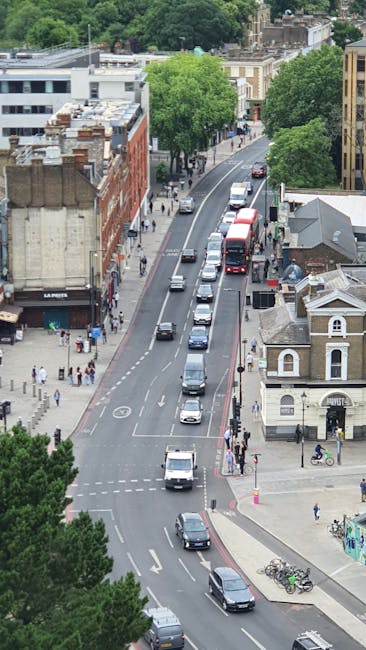An aerial view of a city street during daylight shows a line of vehicles, including vans and cars, moving along a curved road with designated parking spaces. On the left side, pedestrians are walking 