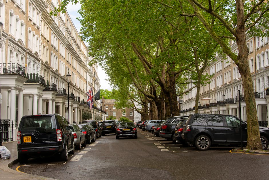 An aerial view of a city street during daylight shows a line of vehicles, including vans and cars, moving along a curved road with designated parking spaces. On the left side, pedestrians are walking 