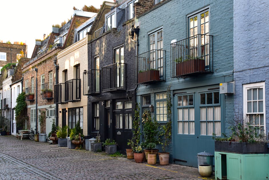 A row of terraced houses on New Kings Road, Fulham, with exterior facades painted in various shades including beige, black, and light blue. Each property has a small balcony with black metal railings and potted plants or flowers. The ground level features a cobbled street with several large potted plants, a bench, and wheelie bins placed outside the front doors. Light from the setting sun casts shadows along the buildings and highlights the textured brickwork and door frames. This scene depicts a typical residential street suitable for house removals or furniture transport services by Fulham Man and Van, illustrating potential challenges for home relocation in narrow access homes while involving loading and unloading activities at street level.