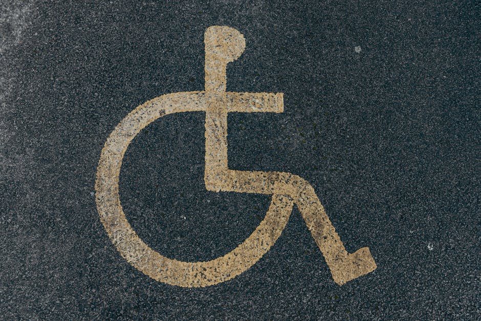 Close-up of a yellow wheelchair accessible parking symbol painted on a rough asphalt surface outside a property, with visible texture and small stones in the pavement. The symbol depicts a simplified outline of a person in a wheelchair, emphasizing accessibility. The background consists of dark grey asphalt with the yellow paint standing out clearly. This image is relevant to the context of house removals and moving services, such as those offered by Fulham Man and Van, especially when considering accessibility for furniture transport, home relocation plans involving narrow or wheelchair-accessible entrances, or parking arrangements for vans during the loading process on New Kings Road.