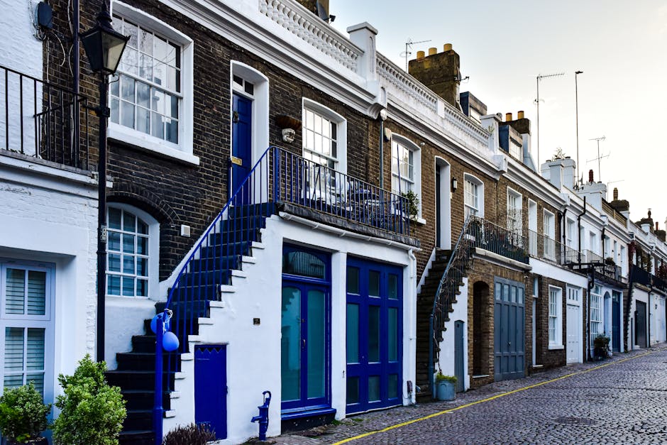 A row of terraced houses on New Kings Road, Fulham, with exterior facades painted in various shades including beige, black, and light blue. Each property has a small balcony with black metal railings and potted plants or flowers. The ground level features a cobbled street with several large potted plants, a bench, and wheelie bins placed outside the front doors. Light from the setting sun casts shadows along the buildings and highlights the textured brickwork and door frames. This scene depicts a typical residential street suitable for house removals or furniture transport services by Fulham Man and Van, illustrating potential challenges for home relocation in narrow access homes while involving loading and unloading activities at street level.
