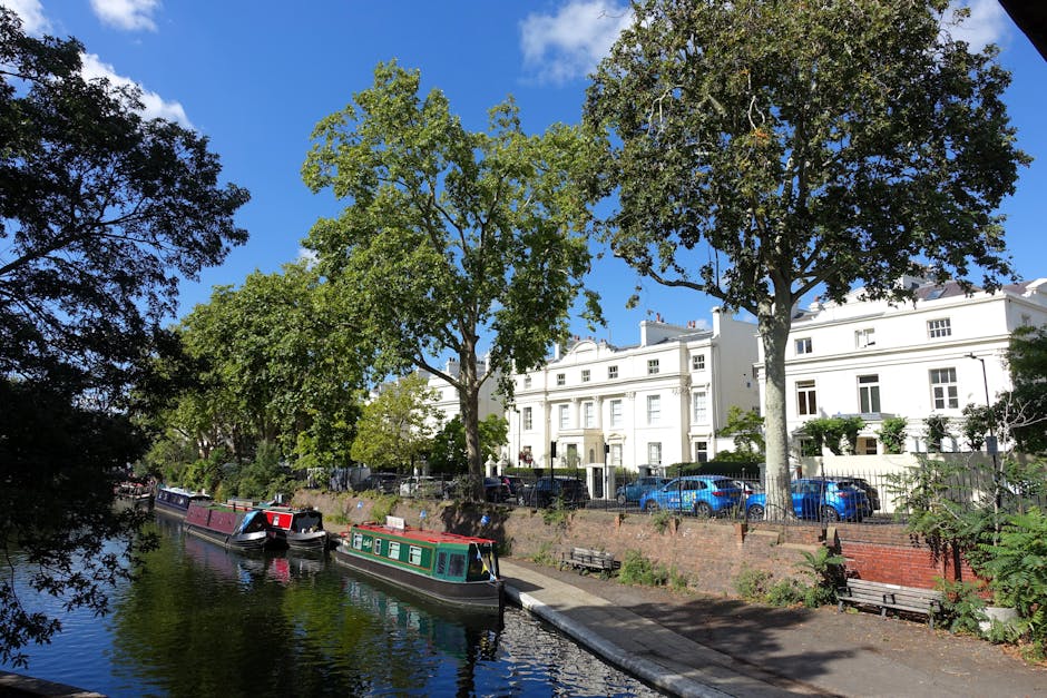 A sunny daytime scene featuring a canal with narrowboats moored along the quay, reflecting the blue sky and surrounding greenery. Tall trees with lush green foliage line the banks, providing shade over benches and a pedestrian walkway. In the background, there are white residential buildings with multiple windows and balconies, some with visible potted plants and decorative elements. Parked cars are seen along the street behind the trees, indicating a residential area. The canal area appears peaceful, with clear weather suitable for home relocation or furniture transport activities. This scene illustrates a typical urban waterways setting in Fulham, which may be relevant for planning house removals or moving services in the area, such as those offered by Fulham Man and Van.