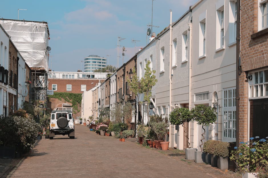 A sunny daytime scene featuring a canal with narrowboats moored along the quay, reflecting the blue sky and surrounding greenery. Tall trees with lush green foliage line the banks, providing shade over benches and a pedestrian walkway. In the background, there are white residential buildings with multiple windows and balconies, some with visible potted plants and decorative elements. Parked cars are seen along the street behind the trees, indicating a residential area. The canal area appears peaceful, with clear weather suitable for home relocation or furniture transport activities. This scene illustrates a typical urban waterways setting in Fulham, which may be relevant for planning house removals or moving services in the area, such as those offered by Fulham Man and Van.